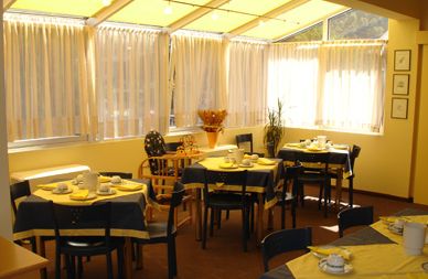 Ein sonnendurchfluteter Essbereich mit mehreren Tischen, die mit gelben und blauen Tischdecken, Tassen, Tellern und Stühlen gedeckt sind, neben großen Fenstern mit durchsichtigen Vorhängen.