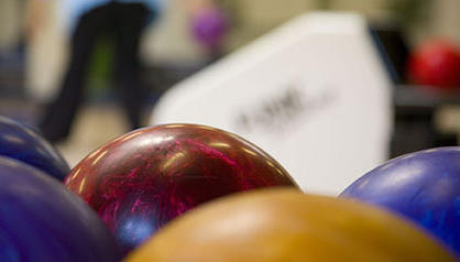 Nahaufnahme von mehreren bunten Bowlingkugeln mit einem unscharfen Hintergrund einer Person und einer Bowlingbahn.