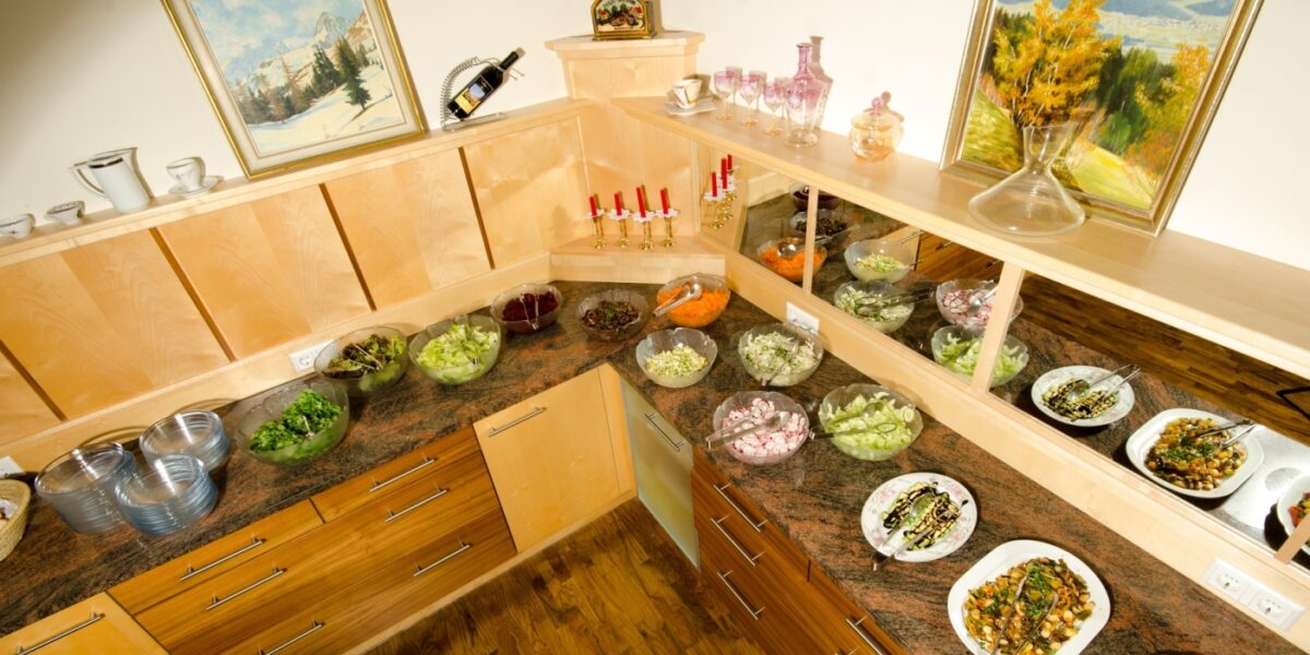 Auf einer Buffet-Theke werden verschiedene Salate und kalte Speisen in Schalen und Tellern angeboten, darüber und an den Wänden stehen Tassen, Kerzen und gerahmte Kunstwerke in Holzregalen.
