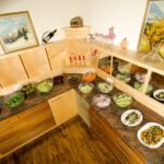 Auf einer Buffet-Theke werden verschiedene Salate und kalte Speisen in Schalen und Tellern angeboten, darüber und an den Wänden stehen Tassen, Kerzen und gerahmte Kunstwerke in Holzregalen.