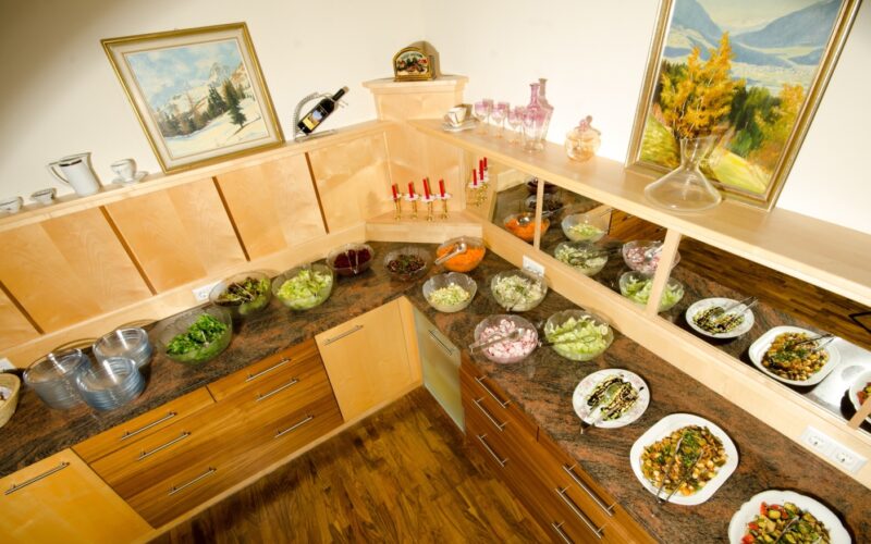 Auf einer Buffet-Theke werden verschiedene Salate und kalte Speisen in Schalen und Tellern angeboten, darüber und an den Wänden stehen Tassen, Kerzen und gerahmte Kunstwerke in Holzregalen.