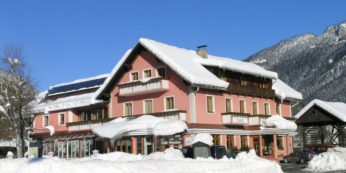 Ein rosafarbenes, mehrstöckiges Gebäude mit schneebedecktem Dach und Umgebung befindet sich in der Nähe einer Straße, mit Bergen und klarem, blauem Himmel im Hintergrund.