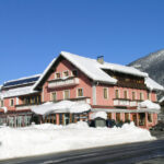 Ein rosafarbenes, mehrstöckiges Gebäude mit schneebedecktem Dach und Umgebung befindet sich in der Nähe einer Straße, mit Bergen und klarem, blauem Himmel im Hintergrund.