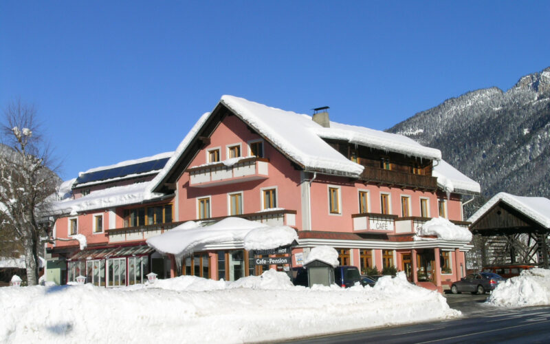 Ein rosafarbenes, mehrstöckiges Gebäude mit schneebedecktem Dach und Umgebung befindet sich in der Nähe einer Straße, mit Bergen und klarem, blauem Himmel im Hintergrund.