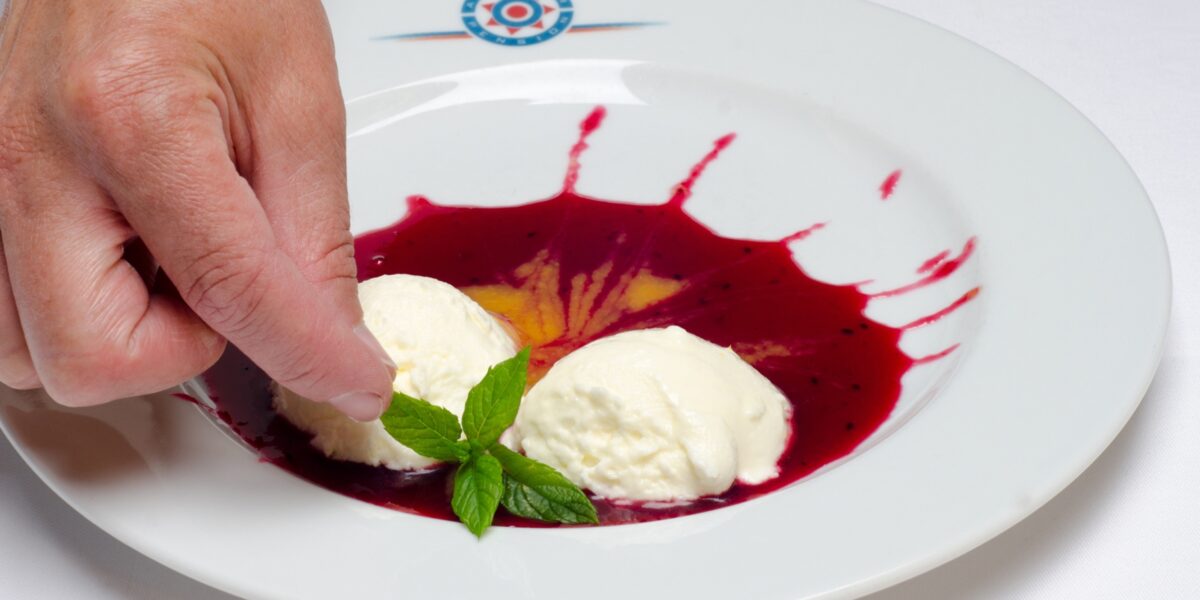Eine Hand platziert einen Minzzweig zwischen zwei Kugeln Vanilleeis auf einem Teller mit roter Beerensauce.