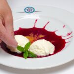 Eine Hand platziert einen Minzzweig zwischen zwei Kugeln Vanilleeis auf einem Teller mit roter Beerensauce.