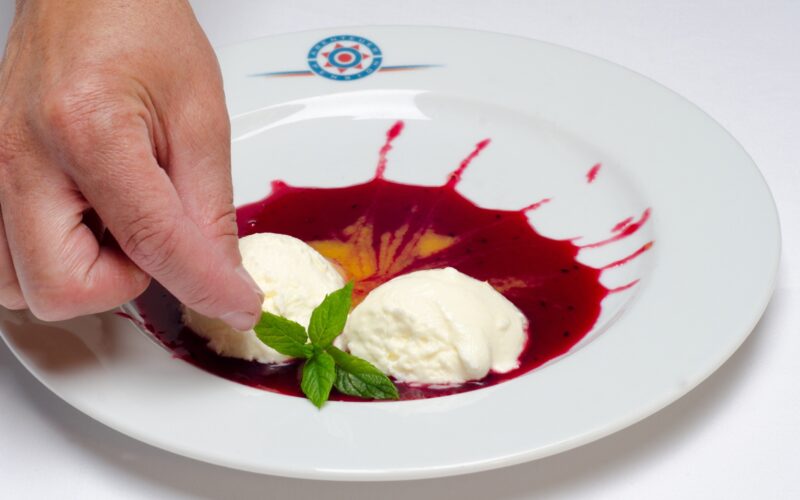 Eine Hand platziert einen Minzzweig zwischen zwei Kugeln Vanilleeis auf einem Teller mit roter Beerensauce.