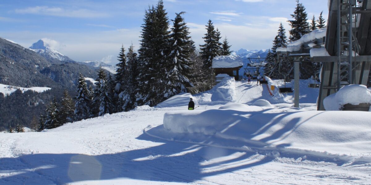 Eine verschneite Gebirgslandschaft mit Bäumen, einem Skilift und zwei Personen, die neben einem Schneehaufen unter einem klaren blauen Himmel stehen.
