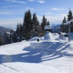 Eine verschneite Gebirgslandschaft mit Bäumen, einem Skilift und zwei Personen, die neben einem Schneehaufen unter einem klaren blauen Himmel stehen.