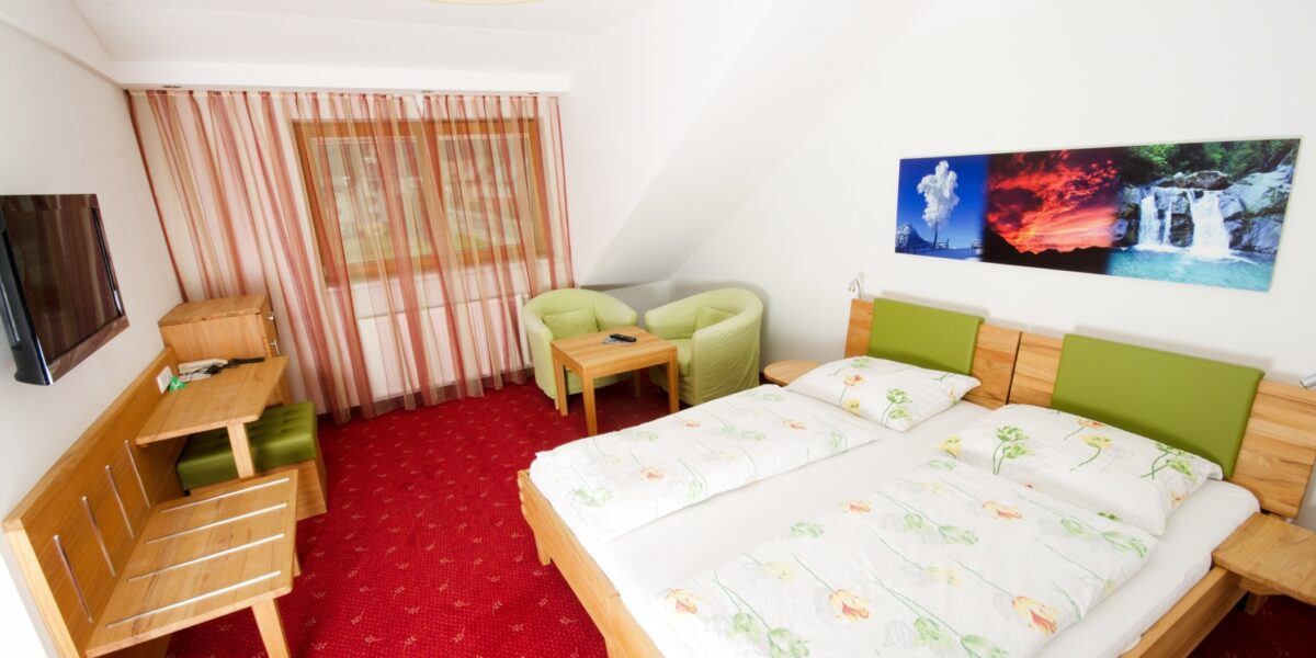 Hotelzimmer mit zwei Einzelbetten, grünen Stühlen, einem Schreibtisch, einem Fernseher und einem Landschaftsfoto an der Wand. Der Boden ist mit rotem Teppich ausgelegt und das Fenster ist mit gestreiften Vorhängen bedeckt.