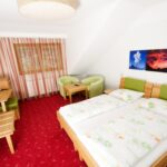 Hotelzimmer mit zwei Einzelbetten, grünen Stühlen, einem Schreibtisch, einem Fernseher und einem Landschaftsfoto an der Wand. Der Boden ist mit rotem Teppich ausgelegt und das Fenster ist mit gestreiften Vorhängen bedeckt.