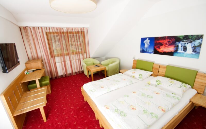 Hotelzimmer mit zwei Einzelbetten, grünen Stühlen, einem Schreibtisch, einem Fernseher und einem Landschaftsfoto an der Wand. Der Boden ist mit rotem Teppich ausgelegt und das Fenster ist mit gestreiften Vorhängen bedeckt.