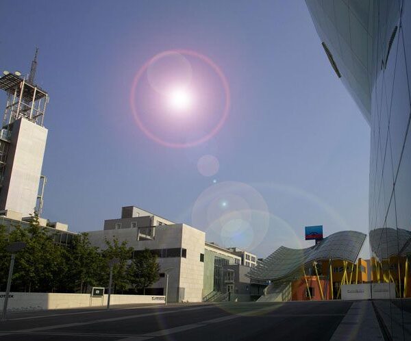 Moderne Stadtstraße mit zeitgenössischen Gebäuden, einem hohen weißen Turm und einer geschwungenen, farbenfrohen Struktur unter einer hellen Sonne mit sichtbarem Linsenreflex am Himmel.