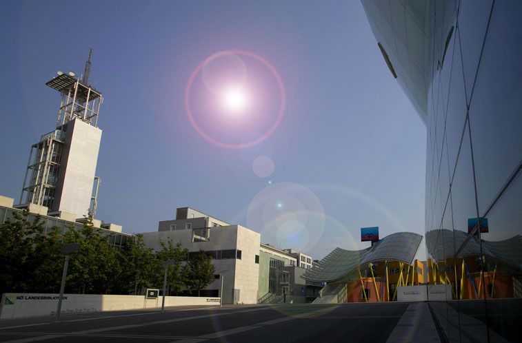 Moderne Stadtstraße mit zeitgenössischen Gebäuden, einem hohen weißen Turm und einer geschwungenen, farbenfrohen Struktur unter einer hellen Sonne mit sichtbarem Linsenreflex am Himmel.