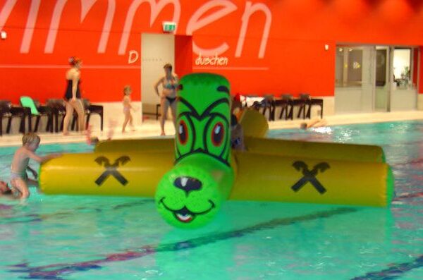 Kinder spielen auf einem großen, grün-gelben aufblasbaren Spielzeug in Form eines Tieres in einem Hallenbad. Im Hintergrund stehen weitere Personen in der Nähe einer roten Wand.