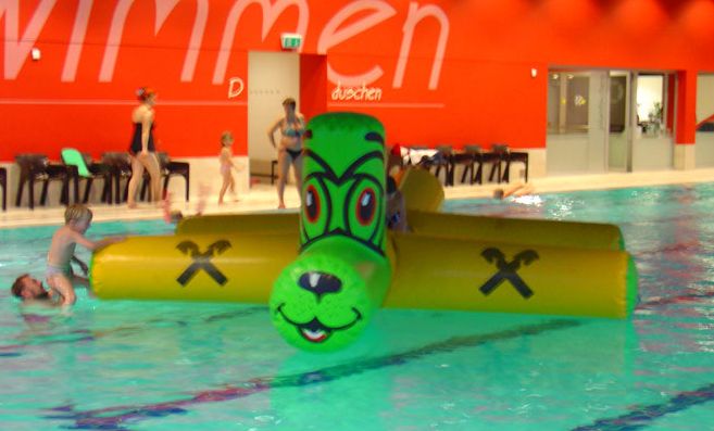 Kinder spielen auf einem großen, grün-gelben aufblasbaren Spielzeug in Form eines Tieres in einem Hallenbad. Im Hintergrund stehen weitere Personen in der Nähe einer roten Wand.