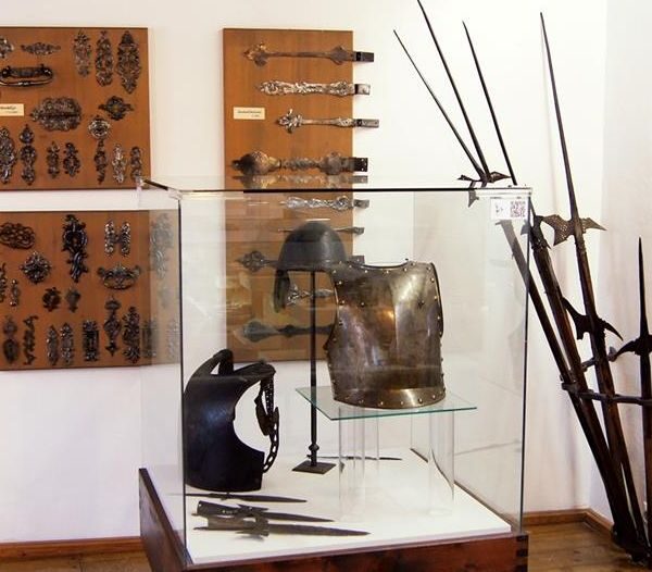 Ausstellung mittelalterlicher Rüstungen und Waffen, darunter Brustpanzer, Helm, Stangenwaffen und dekorative Metallarbeiten, präsentiert in einer Glasvitrine und an den Wänden in einem Museumsambiente.