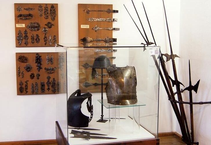 Ausstellung mittelalterlicher Rüstungen und Waffen, darunter Brustpanzer, Helm, Stangenwaffen und dekorative Metallarbeiten, präsentiert in einer Glasvitrine und an den Wänden in einem Museumsambiente.