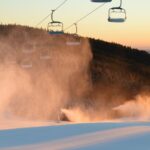 Leere Skiliftsessel ragen bei Sonnenuntergang über einen schneebedeckten Hang, während Beschneiungsanlagen Nebel in die kalte Luft blasen und im Hintergrund bewaldete Hügel zu sehen sind.