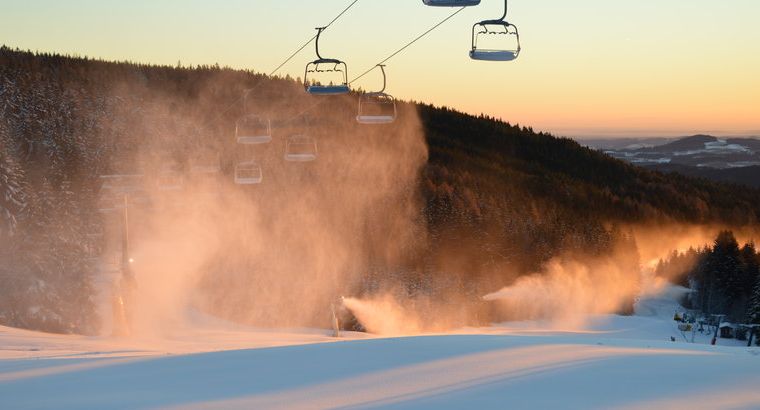 Leere Skiliftsessel ragen bei Sonnenuntergang über einen schneebedeckten Hang, während Beschneiungsanlagen Nebel in die kalte Luft blasen und im Hintergrund bewaldete Hügel zu sehen sind.