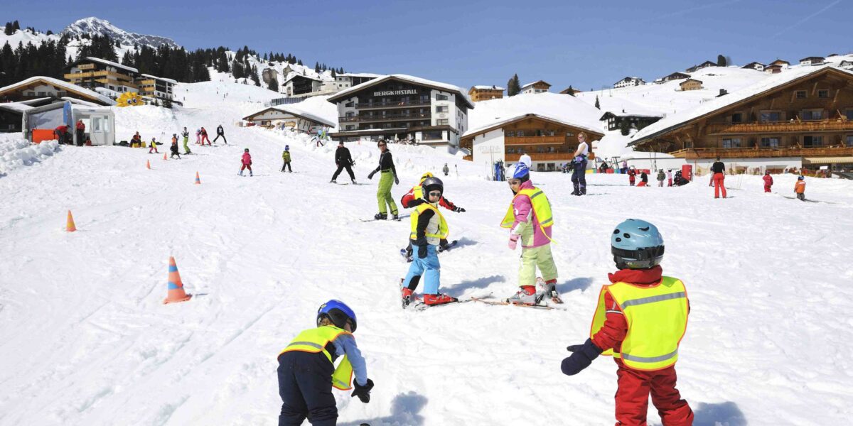 Kinder mit Helmen und Skiausrüstung üben auf einer verschneiten Piste in der Nähe von Holzhütten das Skifahren, während im Hintergrund Erwachsene und andere Skifahrer zu sehen sind.