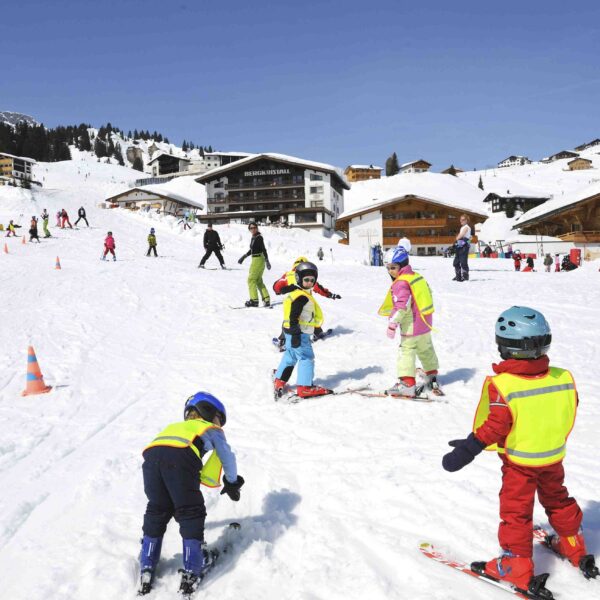 Kinder mit Helmen und Skiausrüstung üben auf einer verschneiten Piste in der Nähe von Holzhütten das Skifahren, während im Hintergrund Erwachsene und andere Skifahrer zu sehen sind.