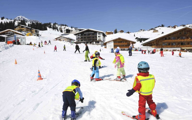Kinder mit Helmen und Skiausrüstung üben auf einer verschneiten Piste in der Nähe von Holzhütten das Skifahren, während im Hintergrund Erwachsene und andere Skifahrer zu sehen sind.