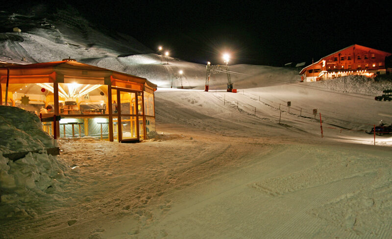 Schneebedecktes Skigebiet bei Nacht mit einem beleuchteten Glasrestaurant und einer Holzhütte, beleuchteten Pisten und sichtbaren Skiliftanlagen.