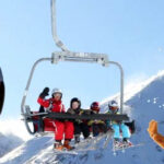 Ein Skilift und eine Gondel befördern Menschen über schneebedeckte Berge, während im Vordergrund eine Person in einem Biber-Maskottchenkostüm winkt.