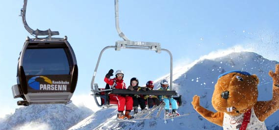 Ein Skilift und eine Gondel befördern Menschen über schneebedeckte Berge, während im Vordergrund eine Person in einem Biber-Maskottchenkostüm winkt.