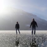 Zwei Personen gehen auf einem zugefrorenen See mit Schneeflecken spazieren, mit einem nebligen Berg und blauem Himmel im Hintergrund.