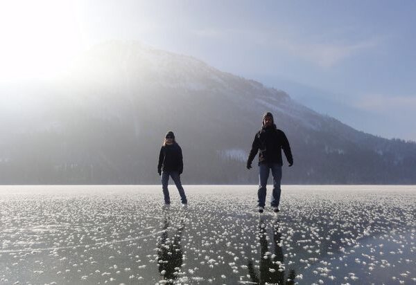 Zwei Personen gehen auf einem zugefrorenen See mit Schneeflecken spazieren, mit einem nebligen Berg und blauem Himmel im Hintergrund.