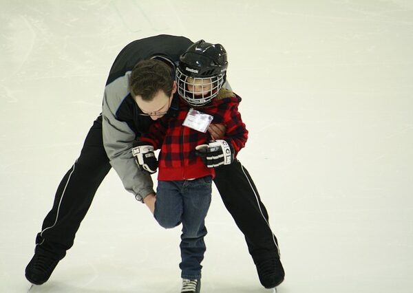 Ein Erwachsener hilft einem Kind, das einen Helm und eine karierte Jacke trägt, auf einer Eislaufbahn aufrecht zu stehen.