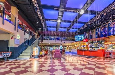 Geräumige Kinolobby mit kariertem Boden, Imbissständen, rot-gelbem Dekor, Filmplakaten, Sitzgelegenheiten im Obergeschoss und beleuchteter Decke.