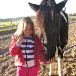 Ein junges Mädchen in einer roten Jacke steht neben einem schwarz-weißen Pferd und streckt dem Pferd in einer sonnigen Umgebung im Freien die Hand zum Schnuppern oder Fressen hin.