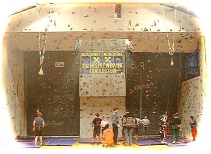 Kinder und Erwachsene stehen und bereiten sich auf das Klettern in einer Kletterhalle mit Kletterwänden und Sicherheitsmatten vor.