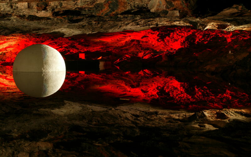 Eine große weiße Kugel schwebt vor einer felsigen, höhlenartigen Oberfläche, die mit rotem Licht beleuchtet ist und Reflexionen nach unten wirft.