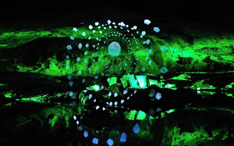 Eine leuchtend grüne Höhlenwand mit einer zentralen kreisförmigen Lichtquelle, umgeben von konzentrischen Ringen aus kleinen blauen Punkten, die ein leuchtendes, abstraktes Muster erzeugen.