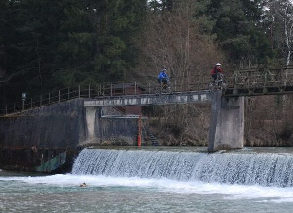 Zwei Radfahrer überqueren eine schmale Brücke über einen Fluss mit einem kleinen Wasserfall. Unten im Wasser nahe der Wasserfallkante ist eine Person zu sehen.