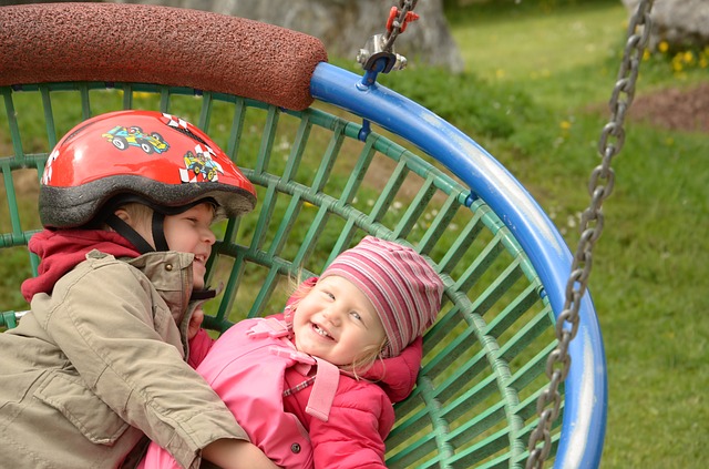 Zwei kleine Kinder, eines davon mit Helm, liegen lächelnd zusammen auf einer großen runden Schaukel auf einem Spielplatz.