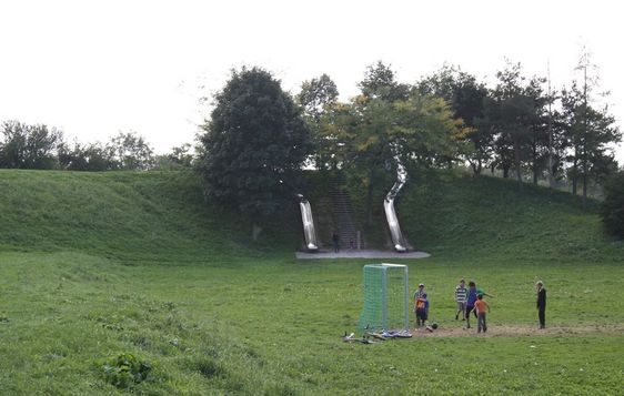 Ein Rasenpark mit zwei Rutschen und Treppen auf einem kleinen Hügel; mehrere Kinder und Erwachsene spielen in der Nähe eines Fußballtors im Vordergrund.