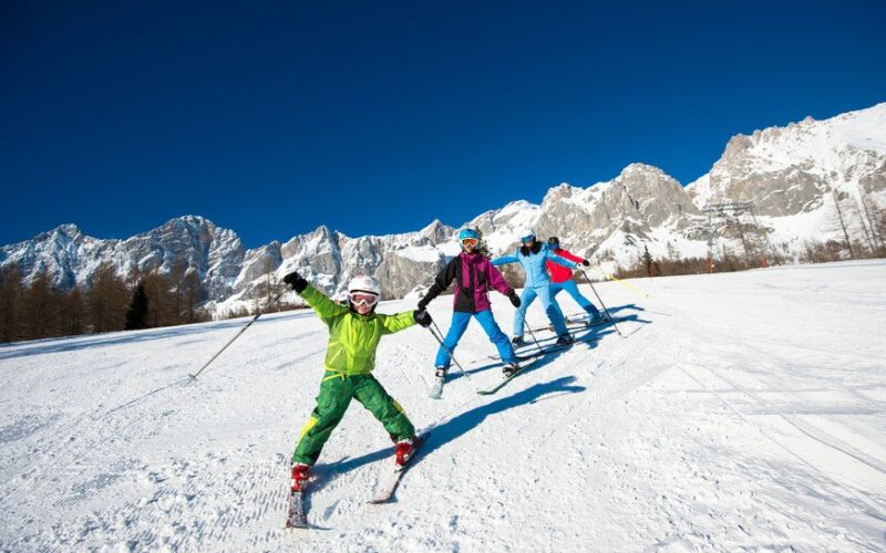 Drei Personen fahren unter einem klaren, blauen Himmel einen schneebedeckten Hang hinunter. Im Hintergrund sind Berge zu sehen. Eine Person vor ihnen breitet freudig die Arme aus.