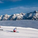 Zwei Skifahrer fahren einen schneebedeckten Hang hinunter, im Hintergrund sind unter einem klaren blauen Himmel eine Bergkette und Wolken zu sehen.