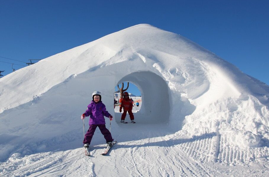 Zwei Kinder in Winterkleidung stehen in der Nähe eines Schneetunnels, in dem sich ein strahlend blauer Himmel und Skispuren befinden.