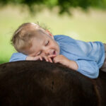 Ein kleines Kind mit hellem Haar und einem blauen Hemd liegt lächelnd und entspannt auf dem Rücken eines dunkelbraunen Pferdes im Freien.