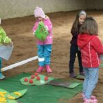 Vier kleine Kinder in bunten Jacken und Mützen spielen drinnen auf einem Sandboden mit Filzmatten und Spielzeugessen.