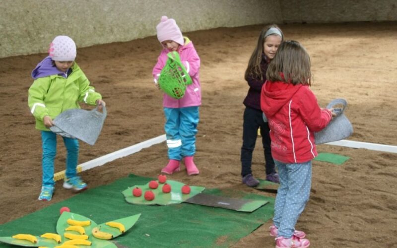 Vier kleine Kinder in bunten Jacken und Mützen spielen drinnen auf einem Sandboden mit Filzmatten und Spielzeugessen.