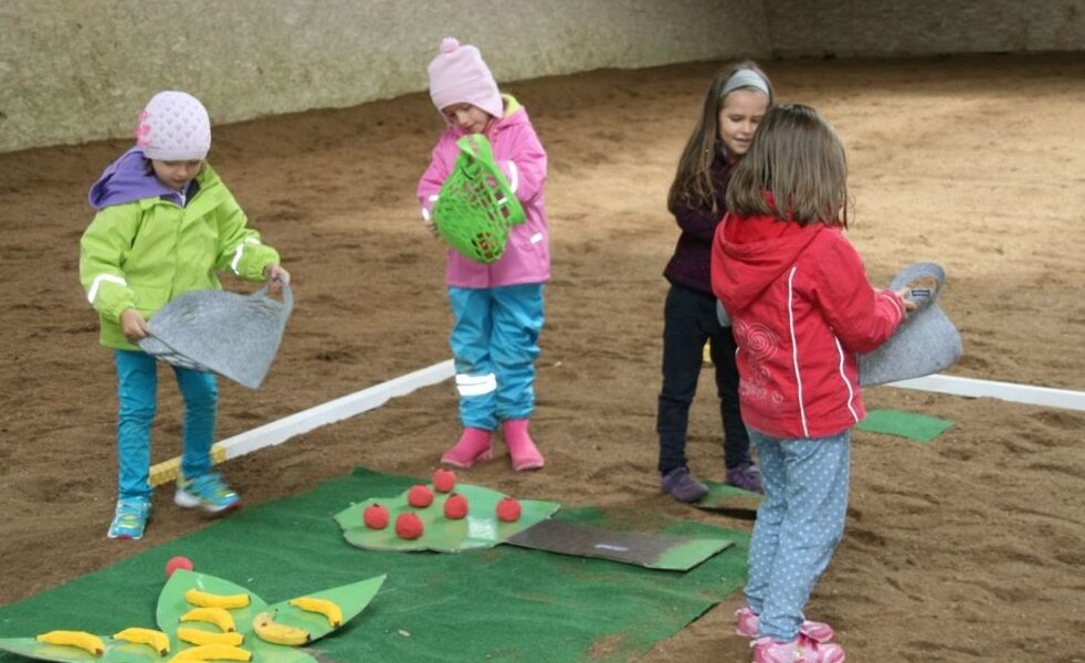 Vier kleine Kinder in bunten Jacken und Mützen spielen drinnen auf einem Sandboden mit Filzmatten und Spielzeugessen.