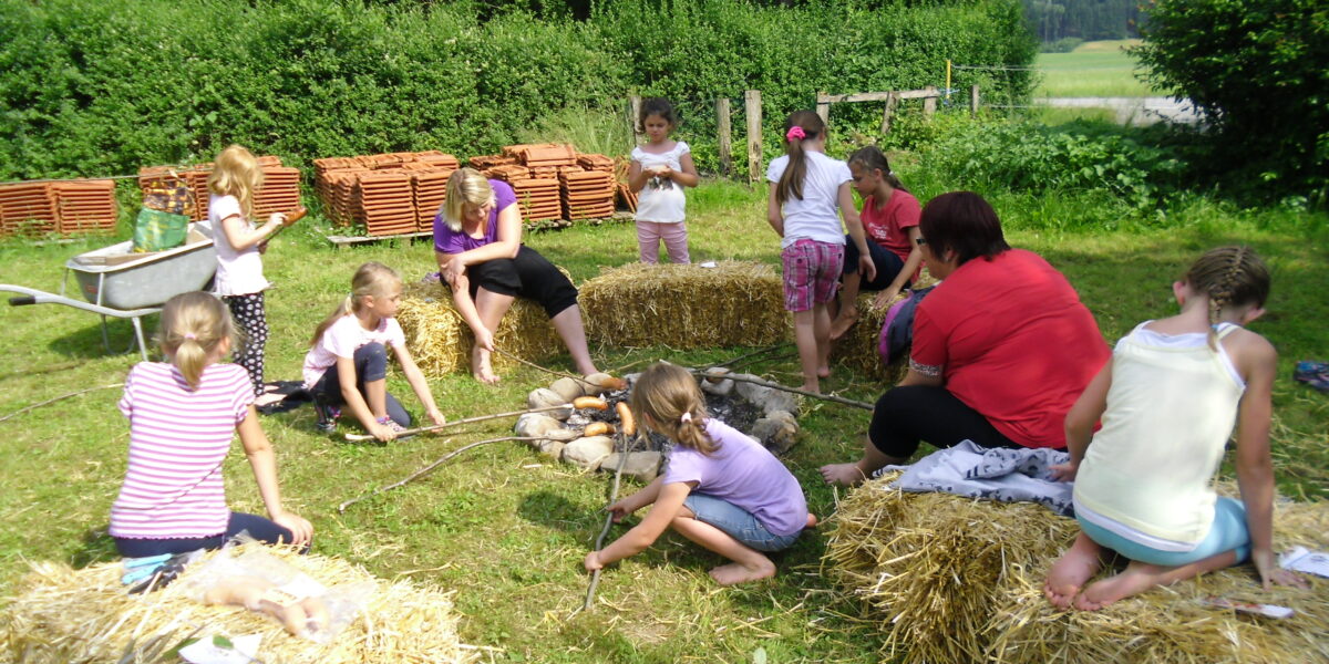 Eine Gruppe von Kindern und Erwachsenen sitzt auf Strohballen um ein Lagerfeuer herum und beschäftigt sich auf einer von Bäumen und Feldern umgebenen Wiese mit Aktivitäten.