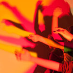 Mehrere Personen heben ihre Arme vor einer Lichtquelle und werfen überlappende, bunte Schatten auf eine Wand.